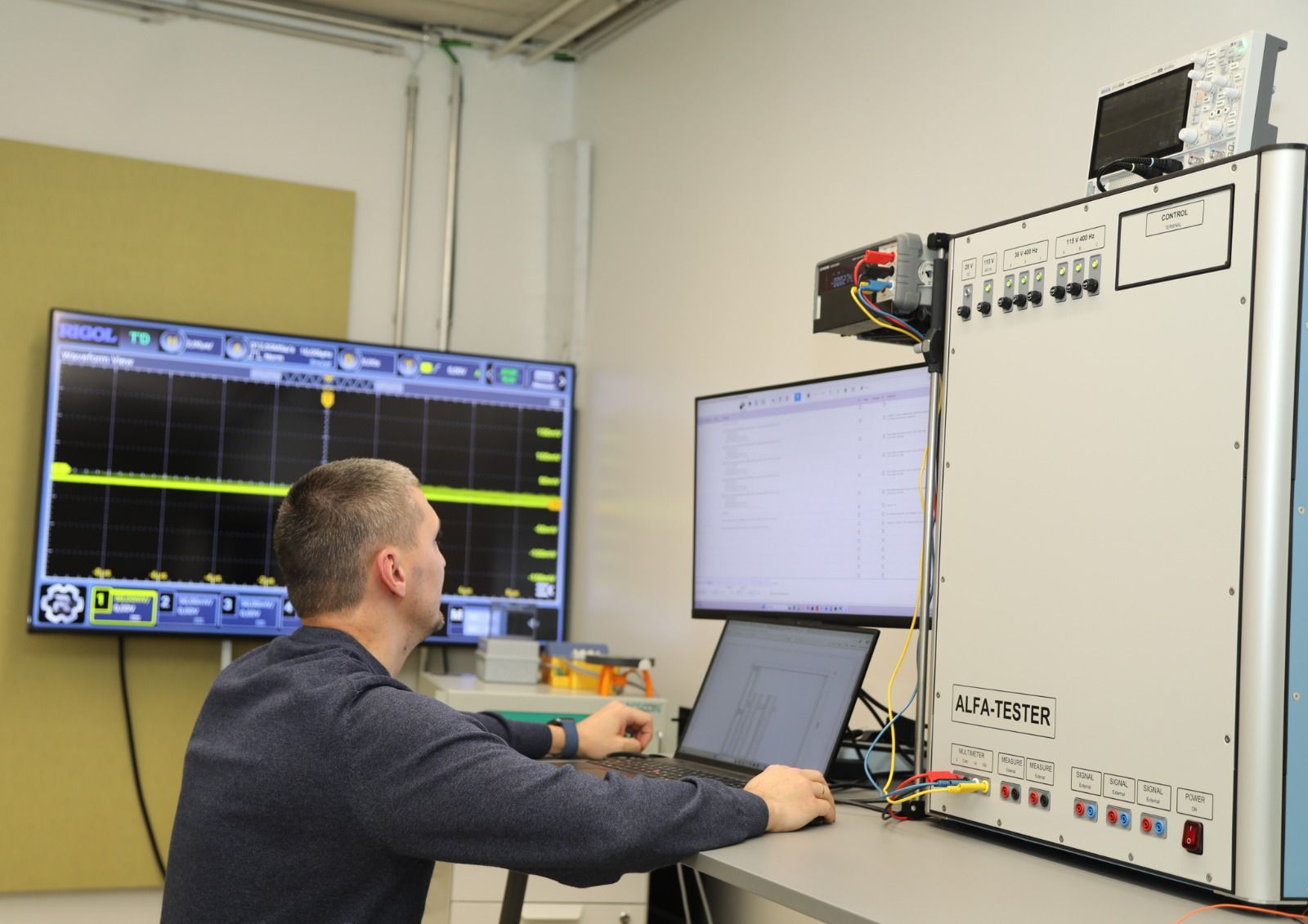 An engineer working at the ALFA-TESTER console, with the test program on a monitor and a waveform on a large screen behind him
